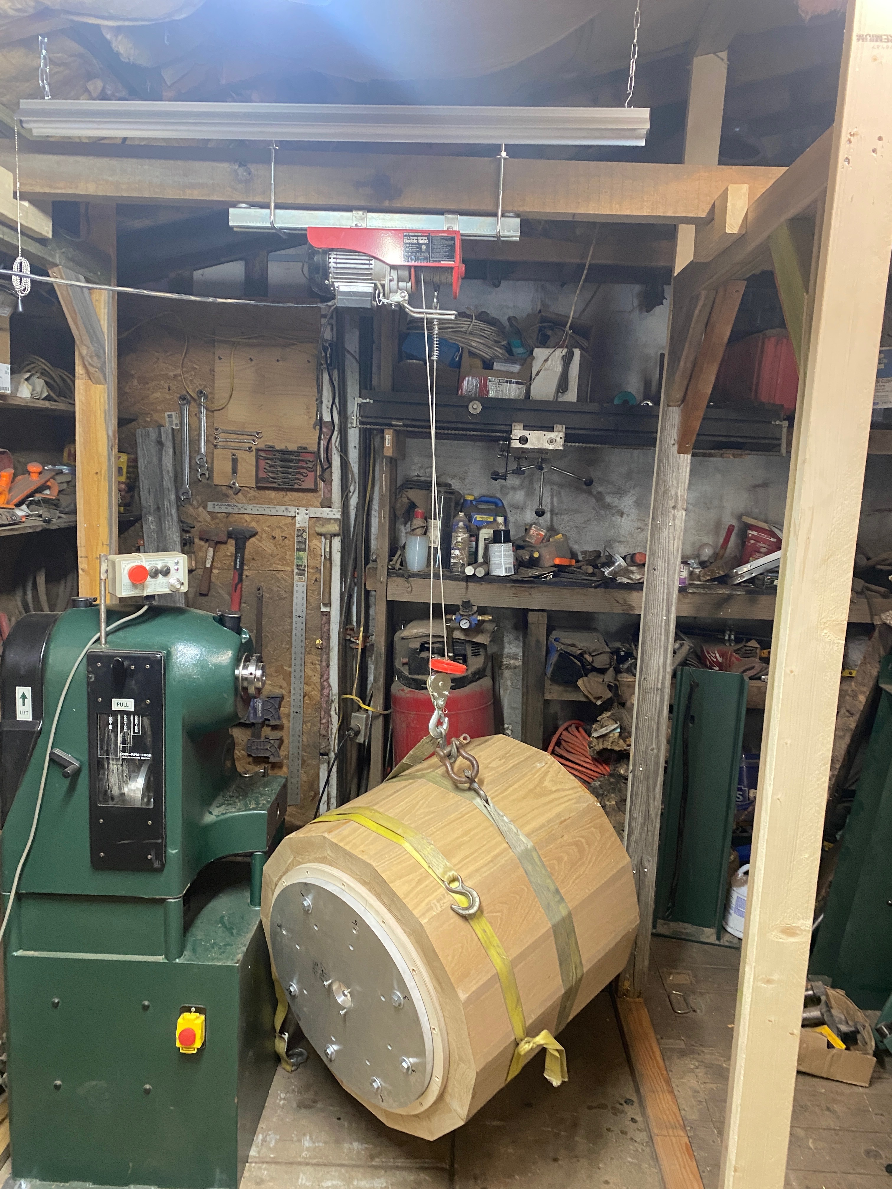 Blank suspended from ceiling hoist on yellow straps, faceplate end toward the lathe headstock.